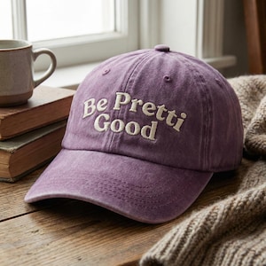 May include: A purple baseball cap with the embroidered text "Be Pretti Good" in white. The cap is made of a soft, textured fabric and has a curved brim. The hat is sitting on a wooden surface.