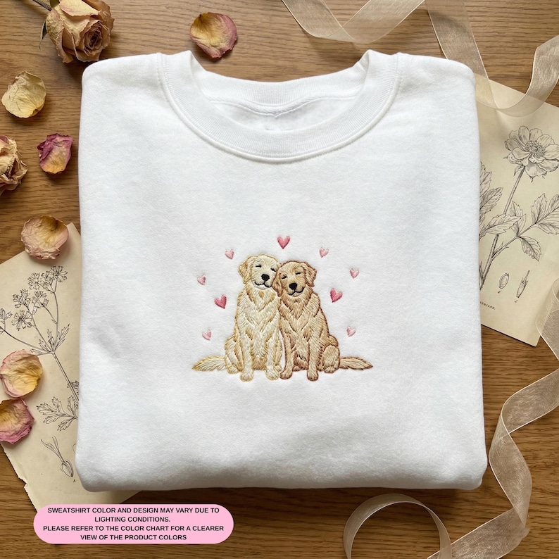 May include: White sweatshirt featuring an embroidered design of two golden retriever dogs with pink hearts. The dogs are sitting close together. The text at the bottom of the image says, "SWEATSHIRT COLOR AND DESIGN MAY VARY DUE TO LIGHTING CONDITIONS. PLEASE REFER TO THE COLOR CHART FOR A CLEARER VIEW OF THE PRODUCT COLORS."