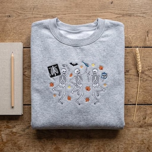 Peut inclure: Un sweat-shirt gris chiné à col rond avec une broderie représentant trois squelettes en train de danser. Les squelettes sont entourés de feuilles d'automne orange, d'une petite chauve-souris noire, d'étoiles jaunes, d'une radio, d'un stylo bleu et d'un presse-papier bleu.
