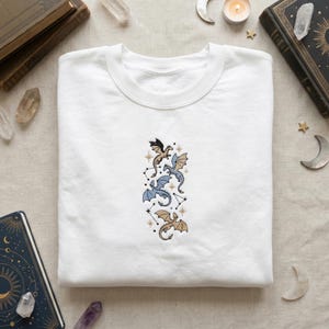 Könnte beinhalten: Weißes Sweatshirt mit Rundhalsausschnitt und einem gestickten Design von drei Drachen in Gold- und Blautönen, mit Sternbilddetails. Die Drachen sind vertikal angeordnet. Das Sweatshirt ist aus weichem Material.