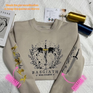 May include: A beige sweatshirt with a black and gold embroidered design of a dragon with a wreath and the text "BASGIATH WAR COLLEGE". The sleeves have embroidered designs, one with a dragon and the text "Fly or die" and the other with a sword and the text "Violent little thing".