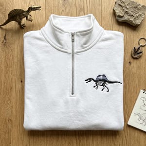 May include: White quarter-zip sweatshirt with a black embroidered Spinosaurus skeleton design. The sweatshirt has a high collar and a silver zipper. The design is on the lower right side of the sweatshirt.