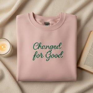 Könnte beinhalten: Hellrosa Sweatshirt mit dem gestickten Text "Changed for Good" in grüner Schrift. Eine brennende Kerze in einem Glas und ein Buch sind ebenfalls im Bild.
