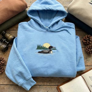 May include: A light blue hoodie with a drawstring hood and a front pocket. The hoodie features an embroidered design of a loon swimming in water with a yellow sun and green trees in the background. Other hoodies are stacked behind it.