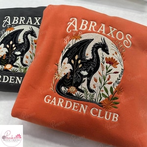 Könnte beinhalten: Zwei Sweatshirts, eines in Schwarz und eines in Orange, jeweils mit einem schwarzen Drachendesign in einem floralen Kranz bestickt. Der Text "ABRAXOS GARDEN CLUB" ist auf das orangefarbene Sweatshirt gestickt. Das schwarze Sweatshirt hat den Text "ABRAX" und "RDEN".