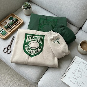 May include: A cream-colored sweatshirt with a green and white "Tommen College" emblem, a green sweatshirt, and a cream-colored sleeve with "Gibsie 7" printed on it. A small succulent plant, scissors, and a wooden tray with embroidery floss are also visible.