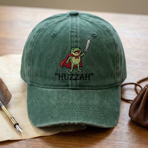 May include: Green baseball cap with a distressed finish. The cap features an embroidered cartoon frog wearing a red cape and holding a sword, with the word "Huzzah" below the frog.