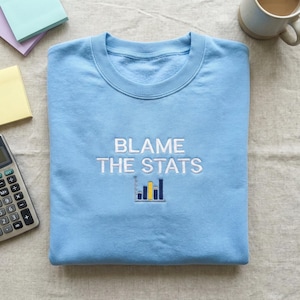 May include: A light blue sweatshirt with the embroidered text "BLAME THE STATS" above a small bar graph. The bar graph has blue and yellow bars. The sweatshirt is folded neatly on a light-colored surface.