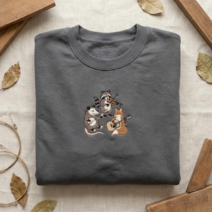 May include: A dark gray sweatshirt featuring an embroidered design of three woodland animals playing musical instruments. A raccoon plays the violin, an opossum the banjo, and a fox the guitar. The sweatshirt is made of a soft material.
