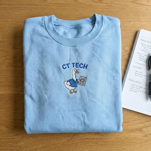 Könnte beinhalten: Hellblaues Sweatshirt mit der gestickten Aufschrift "CT TECH" über einer Cartoon-Gans, die eine blaue Weste trägt und ein Röntgenbild hält. Ein Notizblock und ein Stift sind im Hintergrund zu sehen.
