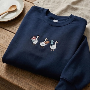 May include: Navy blue sweatshirt featuring embroidered geese carrying a cake, bread, and a whisk. The geese are white with orange feet and beaks. The sweatshirt is made of a soft material and is perfect for anyone who loves baking.