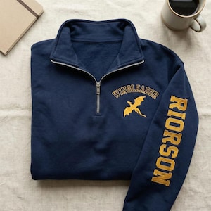 May include: Navy blue quarter-zip sweatshirt with "WINGLEADER" and a gold dragon graphic embroidered on the chest. The right sleeve features "RIORSON" in gold lettering. The sweatshirt is folded on a neutral surface.