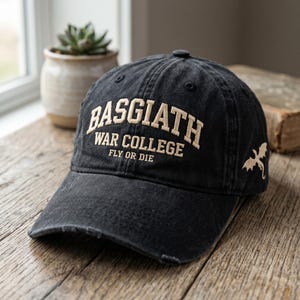 May include: A black baseball cap with a distressed finish featuring the embroidered beige text BASGIATH WAR COLLEGE FLY OR DIE on the front and a small beige dragon silhouette embroidered on the side panel of the cap.