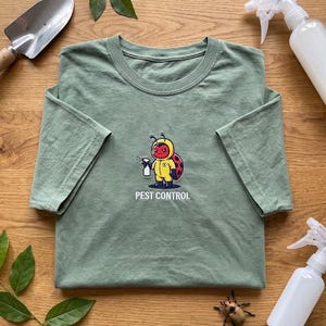 Könnte beinhalten: Ein salbeigrünes Kurzarm-T-Shirt mit einer Grafik eines Marienkäfers in einem gelben Schutzanzug, der eine weiße Sprühflasche hält. Unter der Grafik ist der Text PEST CONTROL in weißen Großbuchstaben auf den Brustbereich gedruckt.