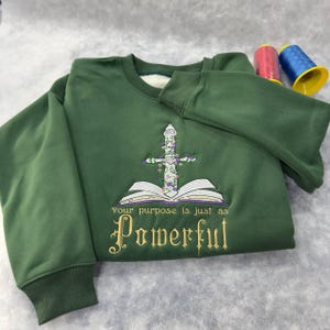 May include: Green sweatshirt with embroidered design. The design features a sword, an open book, and the words "Your purpose is just as Powerful" in gold lettering. The sweatshirt has a crew neck and ribbed cuffs.