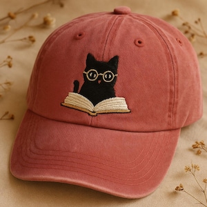 May include: A dusty rose baseball cap featuring an embroidered black cat wearing glasses and reading a book. The cat is the focal point, with the open book below. The cap has a curved brim and an adjustable strap.