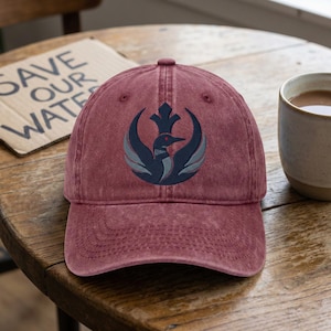 May include: A maroon baseball cap featuring a navy blue and grey bird emblem. The cap has a curved peak and a worn appearance. A cardboard sign with the text "SAVE OUR WATER" is visible in the background.