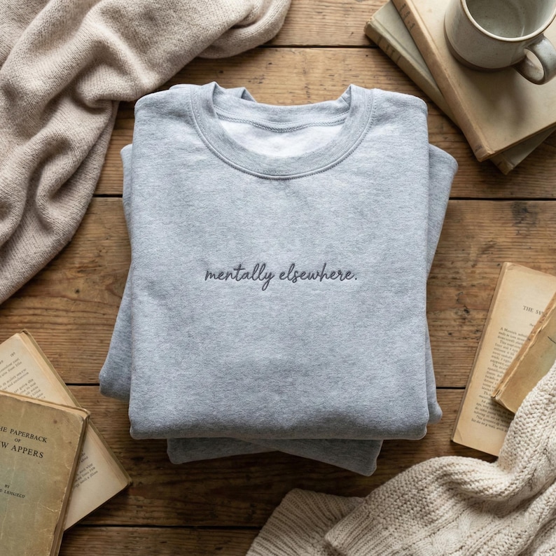 May include: A light gray sweatshirt with the phrase "mentally elsewhere" in black script. The sweatshirt is folded and resting on a wooden surface, surrounded by books and a mug.