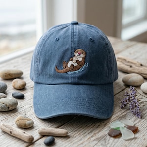 May include: A blue baseball cap with an embroidered otter design. The otter is brown and white, holding a shell while floating in water. The cap is made of a soft fabric and has a curved brim.