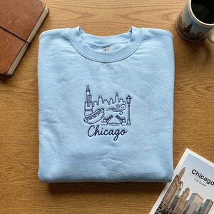 May include: Light blue sweatshirt with a navy blue embroidered Chicago skyline design. The design includes the Willis Tower, a hot dog, a bridge, and the word "Chicago". A mug and a city guide are also visible.