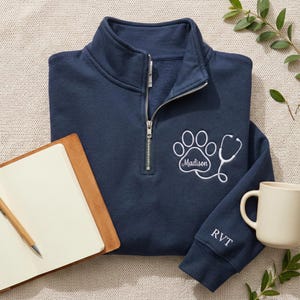 May include: A navy blue quarter-zip sweatshirt featuring a white paw print and stethoscope design. The name "Madison" is embroidered within the paw print. The sleeve has the initials "RVT" embroidered. A notebook and mug are also in the image.
