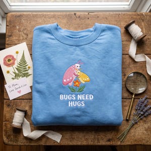 May include: A light blue crewneck sweatshirt featuring an embroidered design of a pink bug and a yellow bug hugging above an orange flower. The text "BUGS NEED HUGS" is embroidered in white below the illustration on the front of the garment.