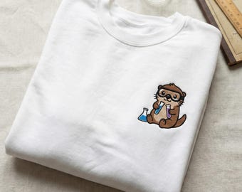 May include: White sweatshirt with an embroidered cartoon otter wearing goggles and holding two beakers with blue and purple liquids. The otter is brown with black accents. The sweatshirt is folded on a neutral surface.