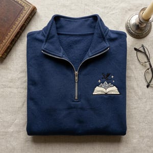 May include: Navy blue quarter-zip sweatshirt with an embroidered design featuring an open book, mountains, a dragon, and stars. The sweatshirt has a silver zipper and a classic collar.