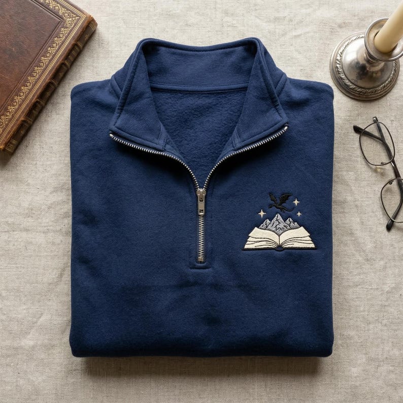 May include: Navy blue quarter-zip sweatshirt with an embroidered design featuring an open book, mountains, a dragon, and stars. The sweatshirt has a silver zipper and a classic collar.