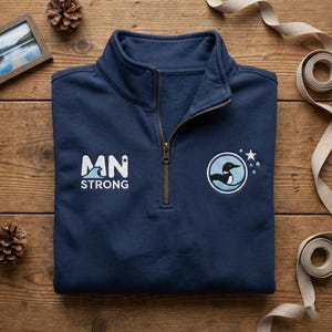 Könnte beinhalten: Ein marineblaues Sweatshirt mit Viertelreißverschluss und dem Text "MN STRONG" in Weiß. Das Sweatshirt hat einen runden Aufnäher mit einem Eisentaucher-Design und einem Stern. Der Reißverschluss hat eine Messingfarbe.