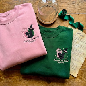 May include: Two crewneck sweatshirts, one pink and one green, with embroidered crests. The crests feature silhouettes, a witch hat, and the text "Changed the World Together." A glass cloche, ribbon, and a "spell page" are also visible.