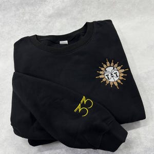 May include: Black crew neck sweatshirt with gold embroidered sun design on the chest and the number "33" on the left sleeve. The sun design has a white face and gold rays.
