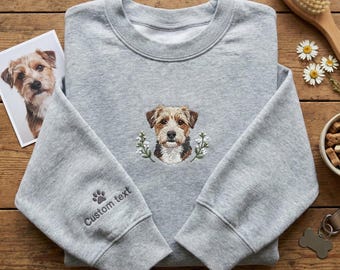 Könnte beinhalten: Ein graues Sweatshirt mit einem gestickten Hundeporträt, umgeben von weißen Blumen. Der Ärmel hat einen Pfotenabdruck und den Text "Custom text". Ein Foto des Hundes liegt neben dem Sweatshirt. Das Sweatshirt liegt auf einer Holzoberfläche.