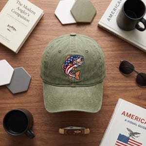 May include: An olive green baseball cap featuring an embroidered graphic of a fish with a red, white, and blue American flag pattern on its body. The cap has a curved brim and a structured crown with stitched eyelets for ventilation.