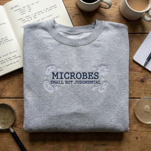 May include: A grey sweatshirt with the navy blue text "MICROBES SMALL BUT JUDGMENTAL". The design includes white embroidered microbe illustrations. The image is set on a wooden surface, with lab notes and coffee cups visible.