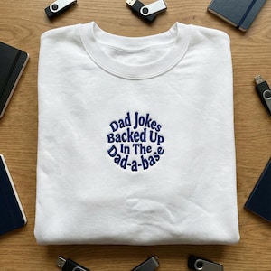 May include: White crew neck sweatshirt with the embroidered text "Dad Jokes Backed Up In The Dad-a-base" in blue. The sweatshirt is surrounded by notebooks and USB drives on a wooden surface.