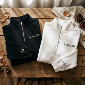 May include: Two personalized quarter-zip sweatshirts, one black and one white, with embroidered text. The black sweatshirt has "J♡A" on the cuff and "I.I.MMXXIV" on the chest. The white sweatshirt has "A♡J" on the cuff and "I.I.MMXXIV" on the chest.