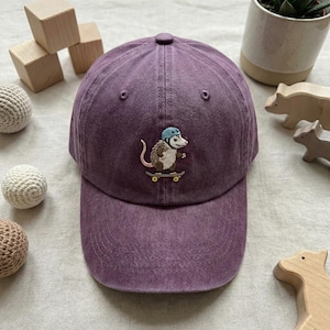 May include: A purple baseball cap featuring an embroidered possum on a skateboard, wearing a blue helmet. The cap is made of a soft, washed fabric with a curved peak. The design is playful and fun.