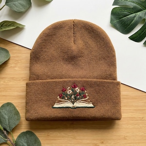 May include: A brown beanie with a folded brim. The beanie features an embroidered design of an open book with red and white flowers and green leaves. The hat is made of a knitted material.