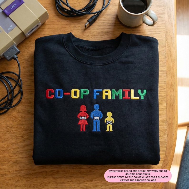 May include: Black sweatshirt with the text "CO-OP FAMILY" in pixelated, colorful letters. Below the text are three pixelated figures: a red figure, a blue figure, and a yellow figure. A coffee mug and a game console are in the background.
