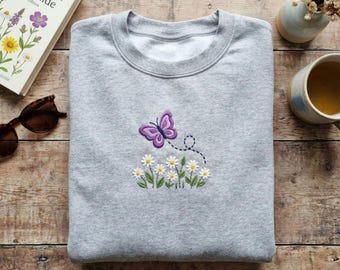 Może przedstawiać: Jasnoszara bluza z haftem przedstawiającym fioletowego motyla i stokrotki. Motyl znajduje się nad rzędem haftowanych stokrotek. Widoczna jest książka zatytułowana "Wildflower Field Guide".