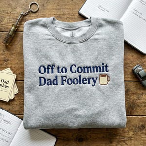 May include: A grey sweatshirt featuring the embroidered text "Off to Commit Dad Foolery" in navy blue, with a coffee cup graphic. The sweatshirt is shown on a wooden surface, alongside other objects.