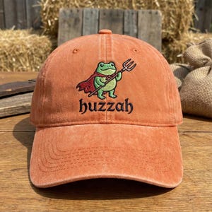 Peut inclure: Casquette de baseball orange avec une broderie d'une grenouille de dessin animé portant une cape et tenant un trident. La grenouille est verte, la cape est rouge et le trident est argenté. Le mot "huzzah" est brodé en dessous en noir.