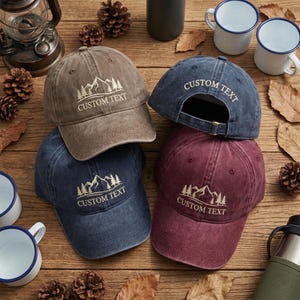 May include: Four baseball caps in brown, dark blue, and maroon, each featuring an embroidered mountain and pine tree graphic with the text CUSTOM TEXT. The caps have a distressed finish, curved brims, and adjustable back straps with metal buckles.