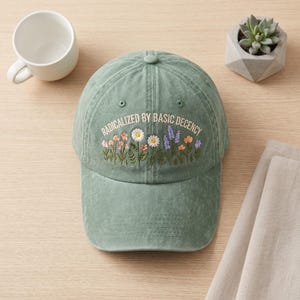 May include: A sage green baseball cap featuring an embroidered design of colourful wildflowers and the text "RADICALIZED BY BASIC DECENCY" in white lettering. The cap has a curved brim, a button on top, and visible stitching across the front panels.