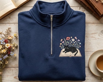 Könnte beinhalten: Marineblaues Sweatshirt mit Viertelreißverschluss und einem gestickten schwarzen Kätzchen auf einem offenen Buch, umgeben von Blumen und Schmetterlingen. Das Sweatshirt hat einen silbernen Reißverschluss und einen gefalteten Kragen.