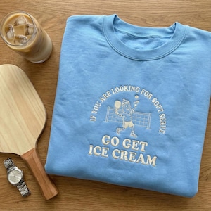 Könnte beinhalten: Hellblaues Sweatshirt mit dem gestickten Text: „IF YOU ARE LOOKING FOR SOFT SERVE GO GET ICE CREAM“. Das Design zeigt eine Cartoon-Gurke beim Pickleballspielen. Ein Holzschläger, eine Uhr und Eiskaffee sind ebenfalls zu sehen.