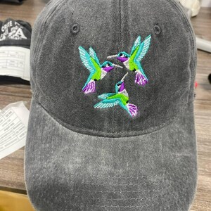 May include: A gray baseball cap with an embroidered design of three hummingbirds. The hummingbirds have teal, purple, and green accents. The cap has a curved brim and a fabric strap closure.