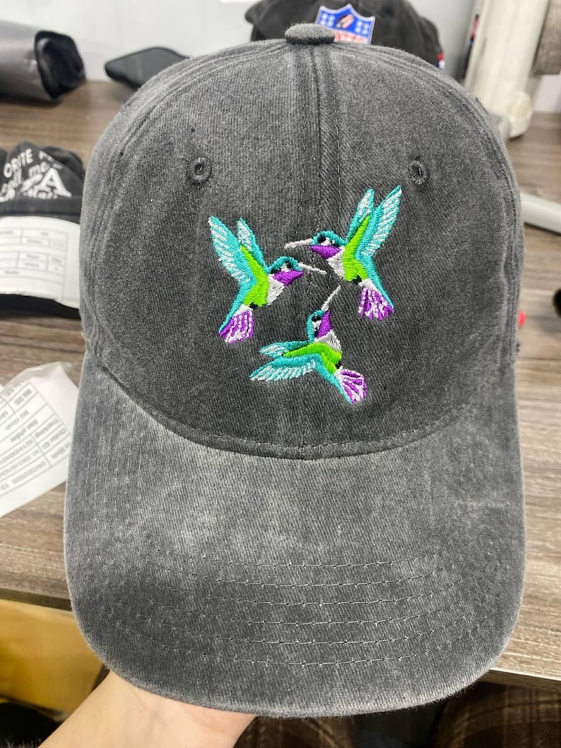 May include: A gray baseball cap with an embroidered design of three hummingbirds. The hummingbirds have teal, purple, and green accents. The cap has a curved brim and a fabric strap closure.