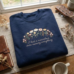 May include: A navy blue sweatshirt with floral embroidery. The design includes various flowers in white, pink, and yellow, with green foliage. The text "I had a marvelous time ruining everything" is embroidered beneath the flowers.